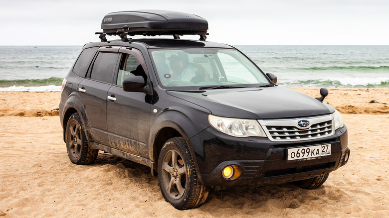 A black Subaru Forester with a cargo box attached on top, at a sandy beach