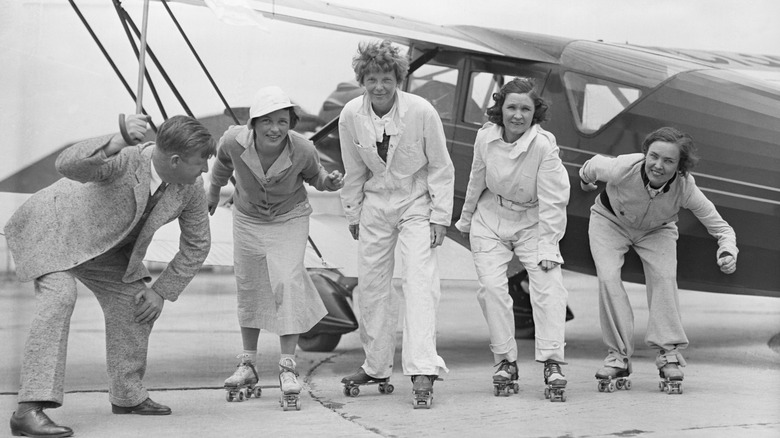 A black and white photo of a bunch of cheerful women wearing roller skates in front of a biplane.