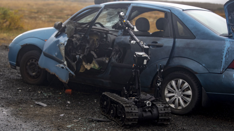 During an training exercise involving improvised explosive devices, a robotic drone examines a bombed-out blue sedan. The driver's seat area has been destroyed.