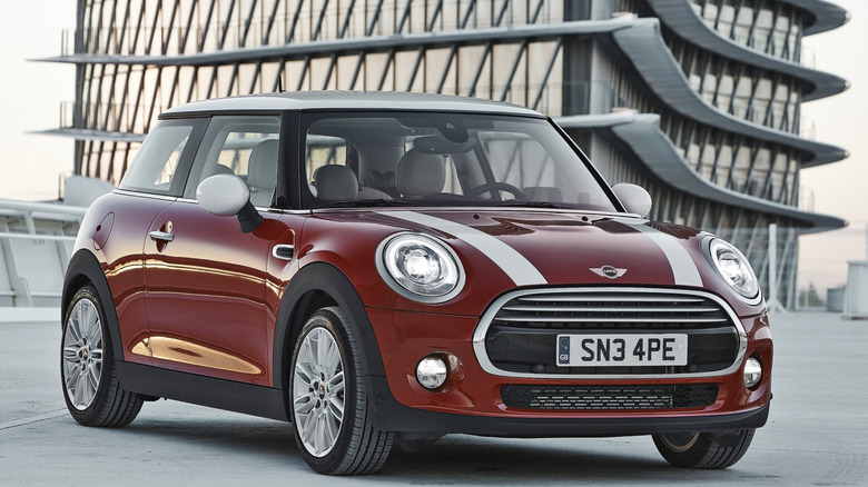 A red mini cooper with white racing strips parked on top of a parking garage, a modernist building in the background