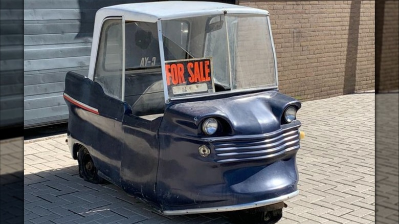 A dark blue, slightly patinaed Electric Shopper on a brick paved parking area alongside a brick building with a steel garage door in teal set behind it.
