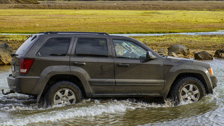 A WK-era Jeep Grand Cherokee drives through deep water with green fields in the background