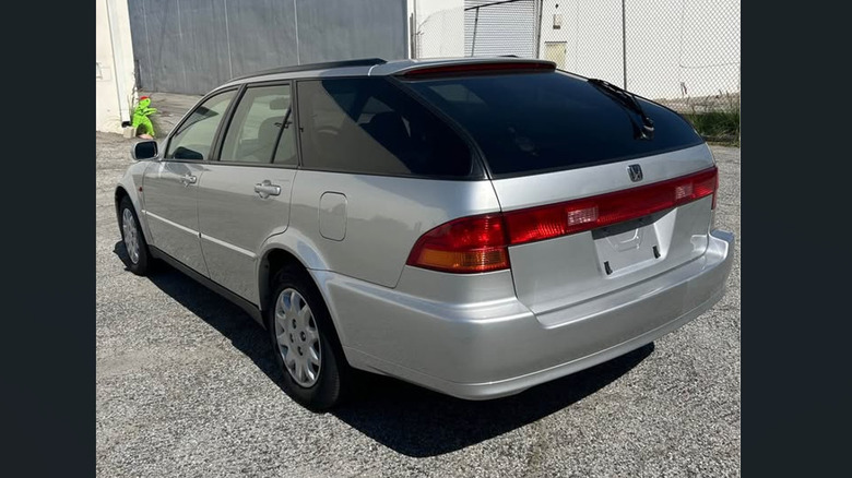 A rear three-quarters shot of a silver 1998 Honda Accord wagon parked on blacktop in front of a yellow cutout of a small person