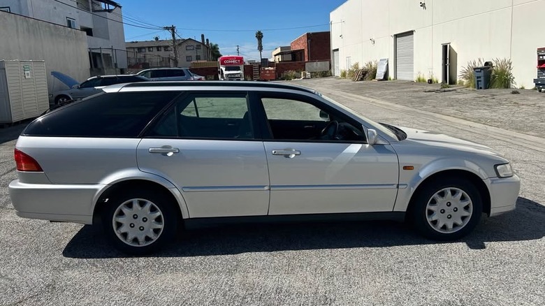 A side view of the silver 1998 Honda Accord wagon parked on blacktop between two white buildings in front of a bus and some plants