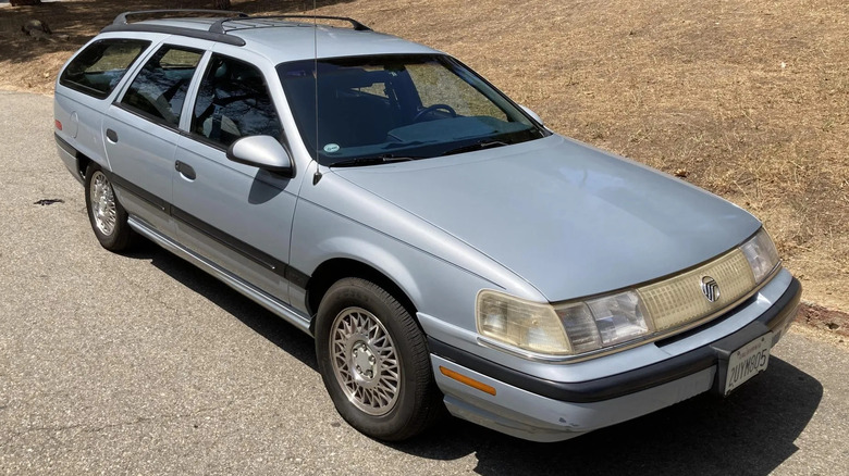 A front three-quarters shot of the light blue Mercury Sable Wagon parked on asphalt next to dead brown grass