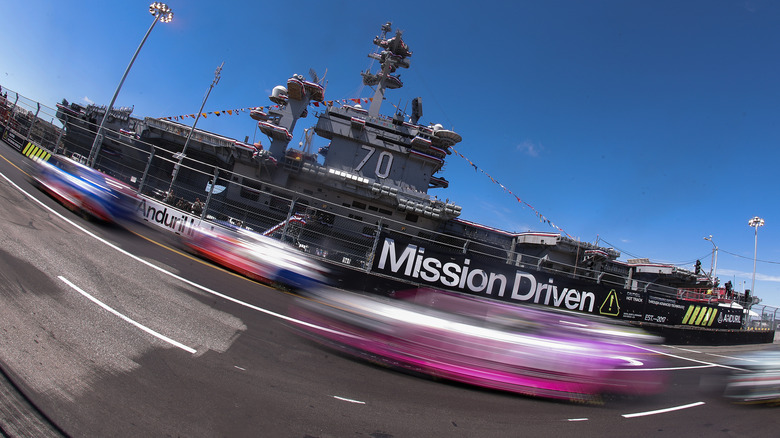 A shot of blurry cars speeding by at a NASCAR race with an aircraft carrier in the background