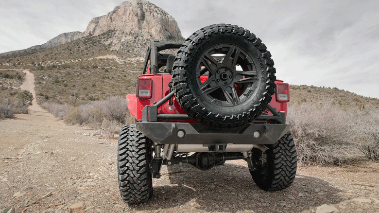 A rear shot of a red Jeep Wrangler parked on a dirt trail in front of a mountain with a large spare Toyo Open Country MT tire on the back