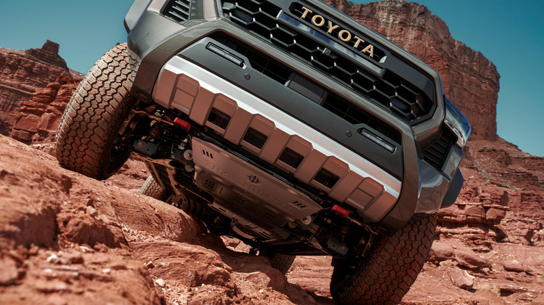A low-angle close-up of a Toyota Tacoma Trailhunter pickup's front end climbing steep red rocks, showing the front skid plate and off-road tires.