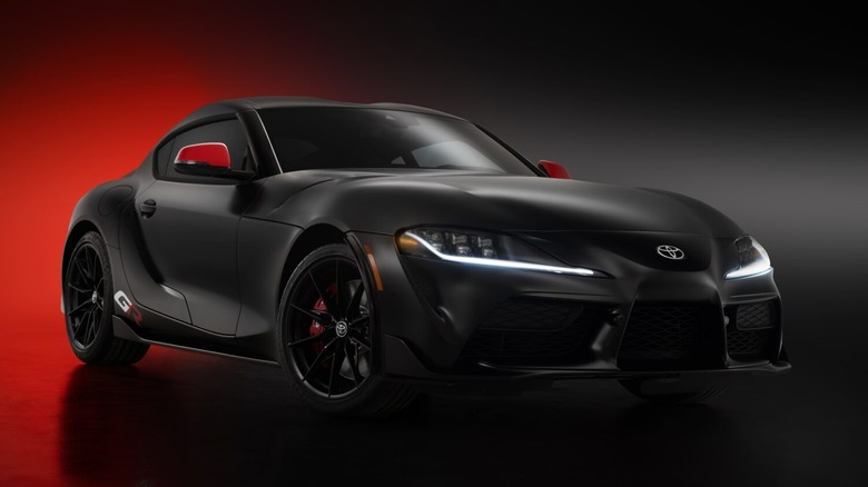 A matte black Toyota GR Supra coupe with red side mirrors illuminated in a dark studio setting.