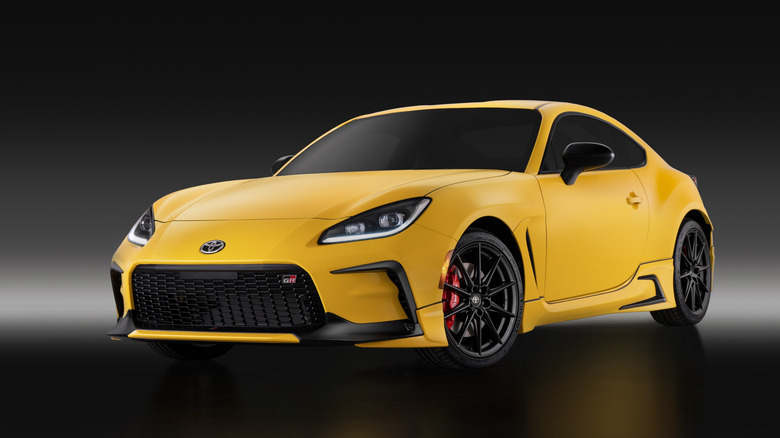 A front three-quarter view of a bright yellow Toyota GR86 sports car with black wheels in a dark studio setting.