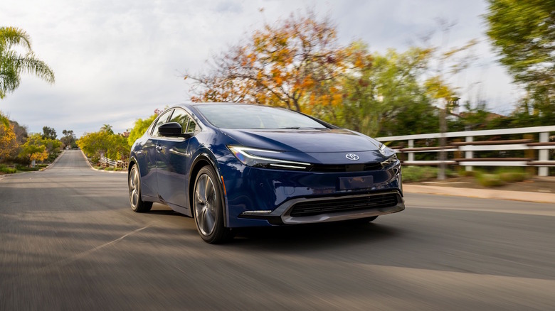 A blue 2026 Toyota Prius on a roadway with a white fence and trees in the background.