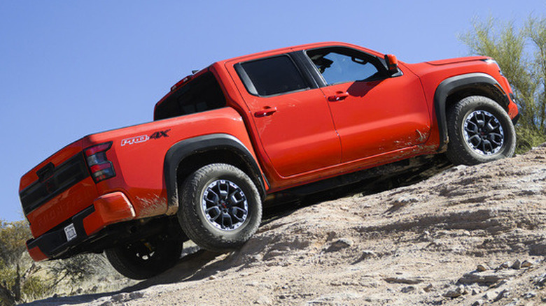 An orange Nissan Frontier PRO-4X pickup truck driving up a steep, sandy and rocky desert hill incline.