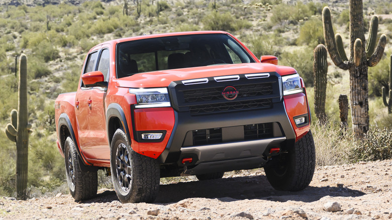 An orange Nissan Frontier PRO-4X crew cab pickup truck parked on a rugged dirt trail amid cacti.