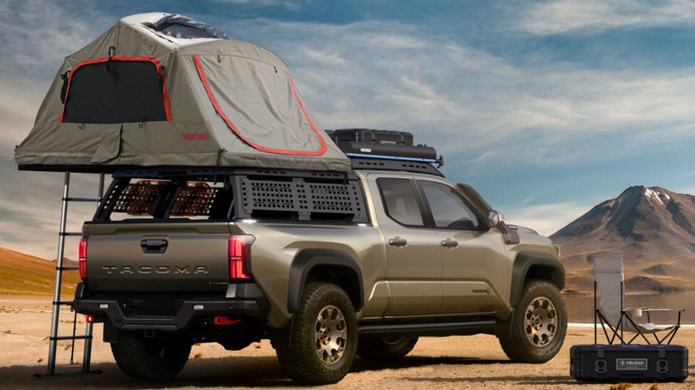 A bronze Toyota Tacoma Trailhunter parked on a sandy beach next to a calm lake, with an arid mountain peak in the background. The truck is heavily upfitted with a bed rack system supporting a deployed Yakima rooftop camping tent.