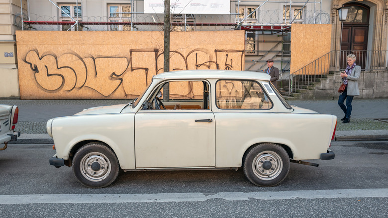 A Trabant car parked near a sidewalk with people walking near it.