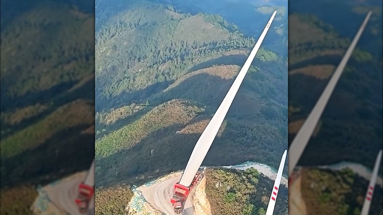 Screenshot of a red small Chinese semi pulling an oversized red semi trailer with a large white blade hanging off of it and the mountain side in China.