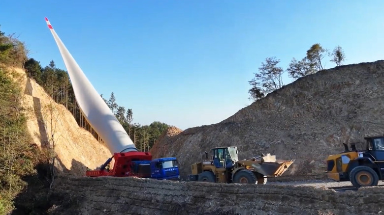 Two yello caterpillar construction diggers lead the way of a small wind turbine hauler with its blade raised at least 60 degrees as it winds a turn on a narrow pass next to a sand dune.