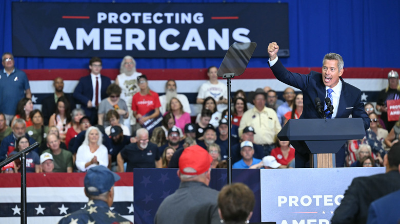 Sean Duffy with his fist in the air, a crowd of americans behind him with a banner that says "protecting americans"