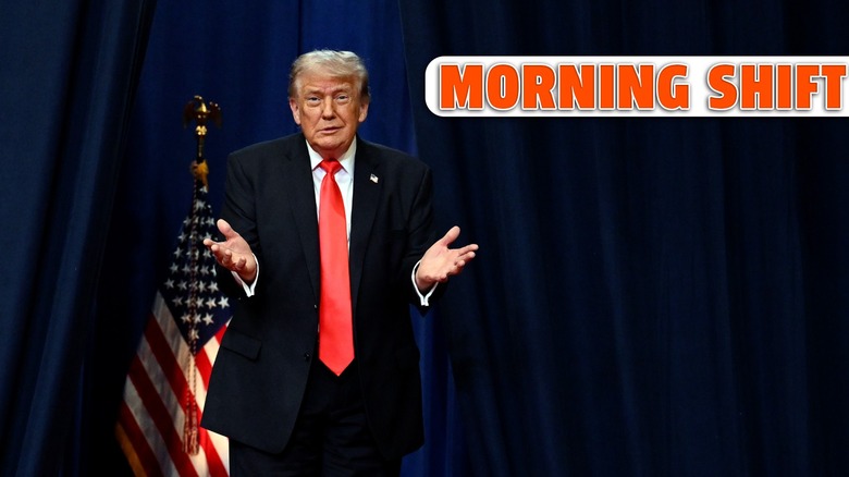 President trump holds his hands out while standing in front of a blue curtain and a single american flag