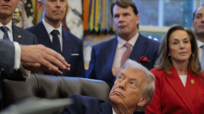 President trump looking up as a hand come from the left side of the image. Behind him is Ford CEO Jim Farley and several other people.