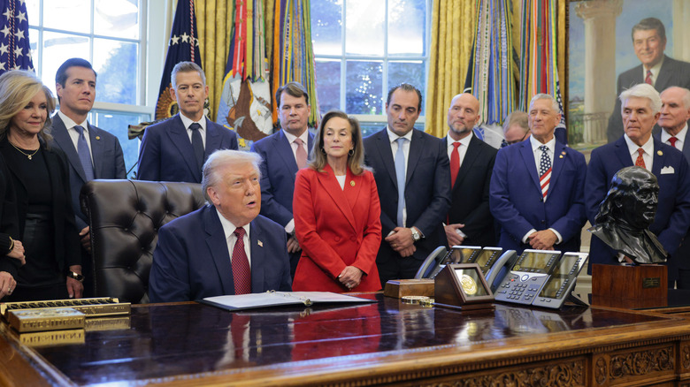 President trump at his desk in the oval office with a bunch of people standing behind him