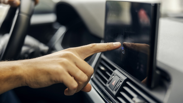 Person's hand interacting with a modern in-car touchscreen display, selecting an option on the control panel for navigation and infotainment features while driving