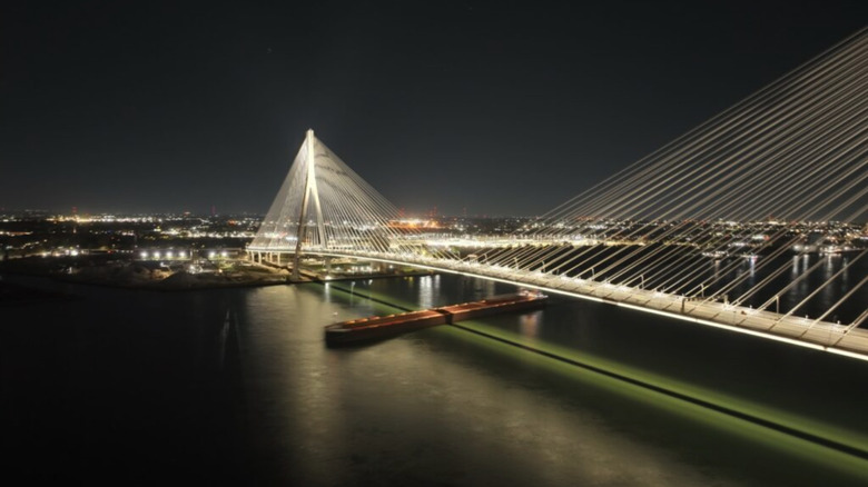 A large barge passes under the illuminated Gordie Howe Bridge on the Detroit River at night.
