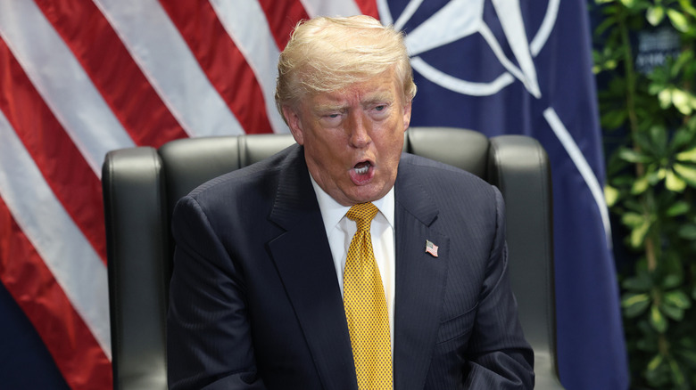 President Trump possibly mid-sentence at a conference, sitting in a chair with the American flag behind him