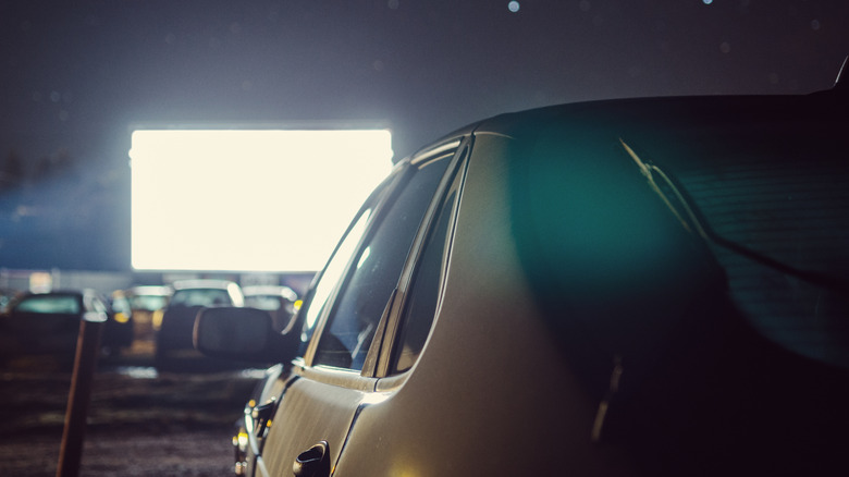 A gold brown older four-door sedan is parked in front of an illuminated screen at a drive-in theater.