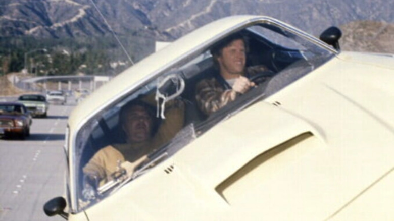 Two men in a yellow car that's tilted sideways driving along a mountainous highway, a scene from "The Gumball Rally"