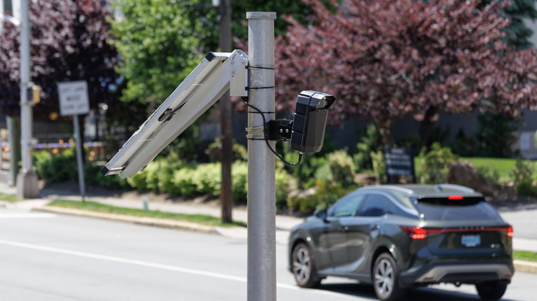 A solar-powered ALPR is angled towards traffic on a small town road. A crossover is braking in the background.