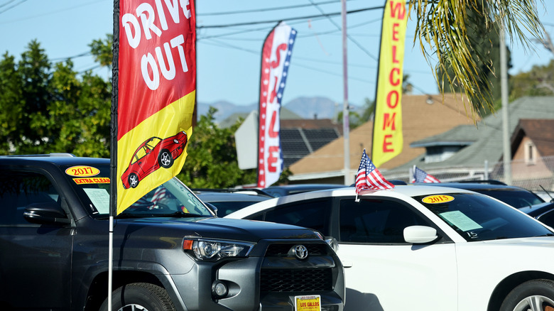 A used car lot with a used grey Toyota 4-Runner, a white Dodge Charger, and other vehicles, with sales and welcome flags waving amid the cars.