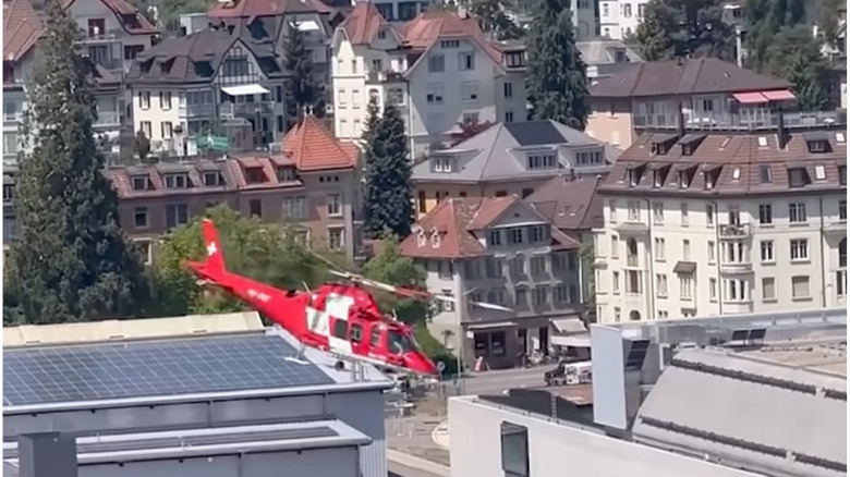 A screenshot showing the red and white Swiss air ambulance flying sideways in front of several multi-story buildings on its approach