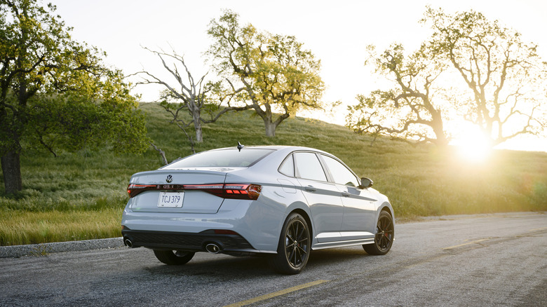 Rear 3/4 view of a gray Volkswagen Jetta GLI driving off into the sunset with a grassy hill and trees in the background