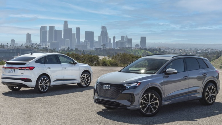 Front three-quarters view of a gray Q4 E-Tron in front of a rear three-quarters view of a white Q4 E-Tron Sportback parked on asphalt in front of downtown LA