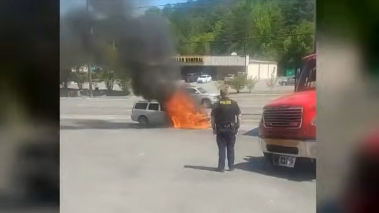A screenshot from the news broadcast showing cell phone video of the white Volvo V70 with the whole hood in flames and a police officer staring at it