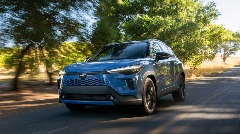 A Cavalry Blue 2026 Toyota Corolla Cross Hybrid XSE driving through a sun-drenched wooded area.