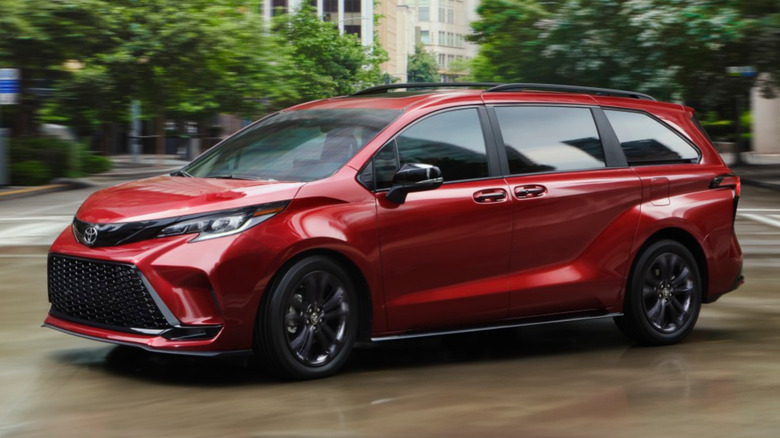 A bright red 2026 Toyota Sienna XSE minivan driving through a modern downtown area with heavy motion blur on the surrounding city trees.