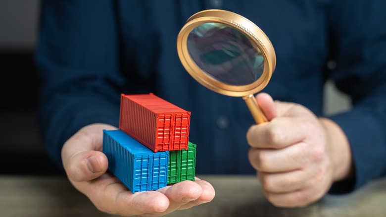 An abstract image of an individual holding a magnifying glass and looking at small different colored shipping containers.