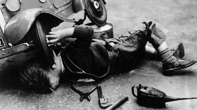 a little boy works under his toy car to "repair" it. The image is black and white and from the 1930s