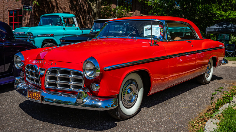 A reddish 1955 Chrysler C-300 sits by trees astatine a car show. A achromatic Ford two-door coupe sits adjacent to it and a bluish Chevrolet pickup motortruck is down it.