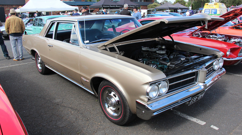 A champagne-colored 1964 Pontiac GTO with its hood open reveals its 389 Tri-Power V8, sitting at a car show next to a red 1960s Ford Mustang