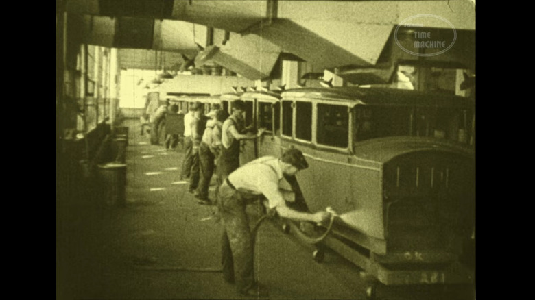 The sepia-toned image of men painting and prepping cars on an old car factory line.