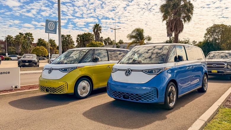 two VW ID Buzz vehicles, one yellow one blue, parked at a VW dealership on a sunny day in California. There are palm trees in the background and white clouds in the sky