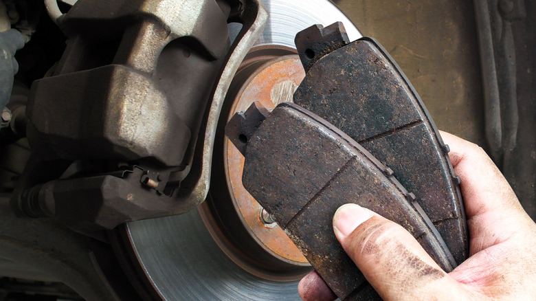 A person holding two brake pads in front of a car's braking assembly.