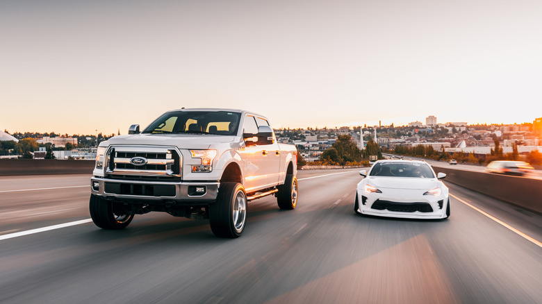 A white Ford truck driving alongside a tuned white Subaru BRZ on the highway at sunset.