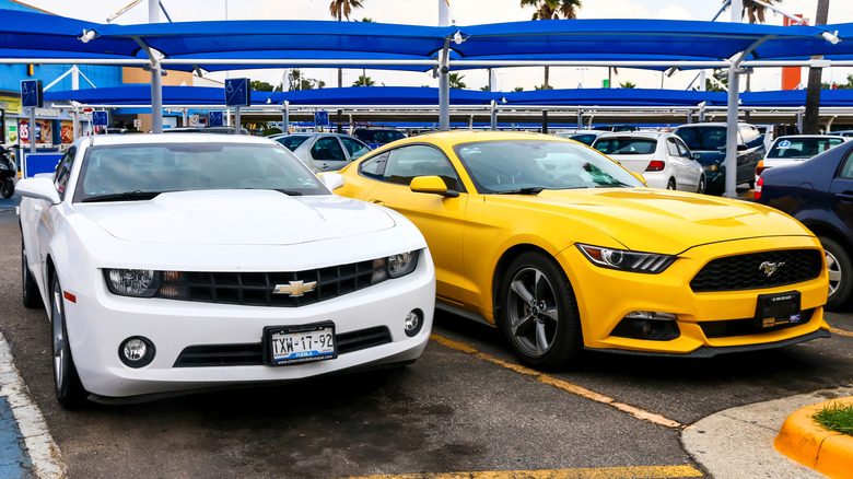 A side-by-side of a white Chevrolet Camaro and a yellow Ford Mustang in a car park.