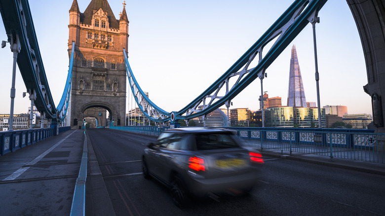 A blurry Mini drives across the London Bridge with city buildings in the background