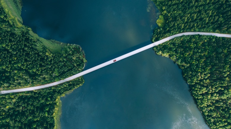 A red car driving over a bridge in Finland with water and green trees surrounding the road