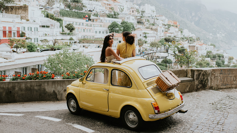 A yellow Fiat Topolino with two women standing through the sunroof parked on cobblestone in front of a concrete wall overlooking old, gorgeous Italian homes on a mountainside
